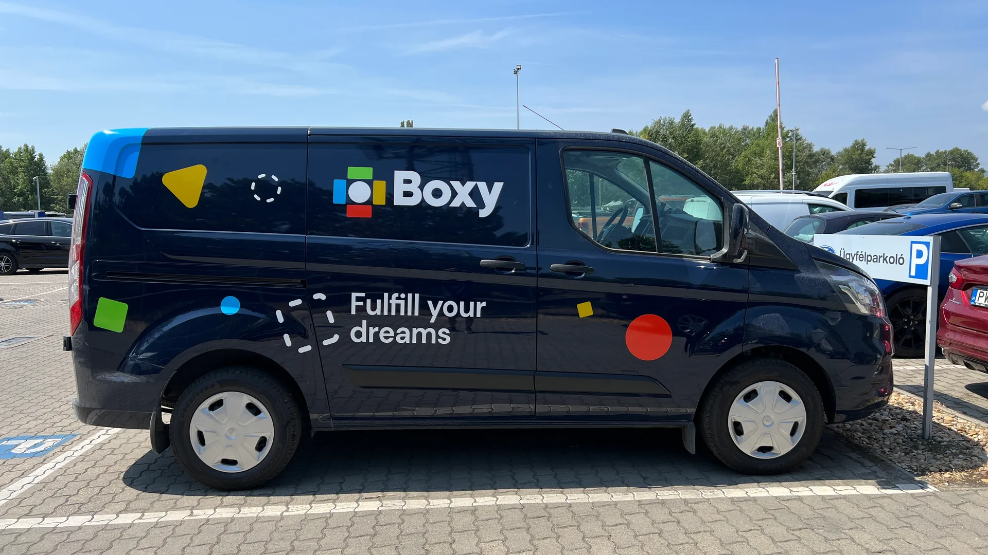 Boxy Ford Transit Custom oldalnézet napsütésben – sötétkék karosszéria, fehér Boxy logó és Fulfill your dreams tagline, élénk színes geometrikus formák