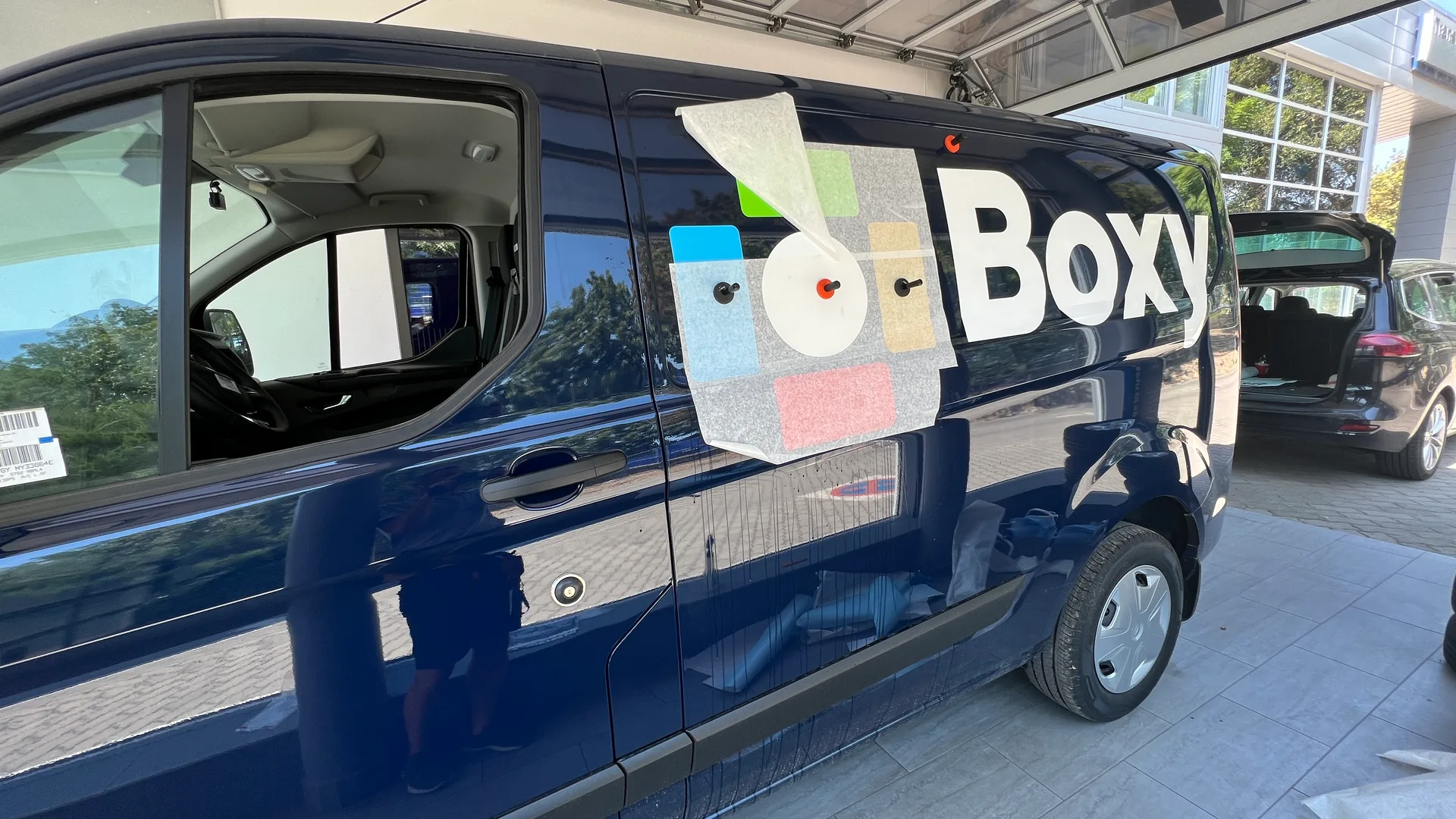 Boxy Ford Transit Custom oldal-dekor telepítés közben – applikációs fólia rajta a Boxy logóval, geometrikus elemekkel a sötétkék karosszérián