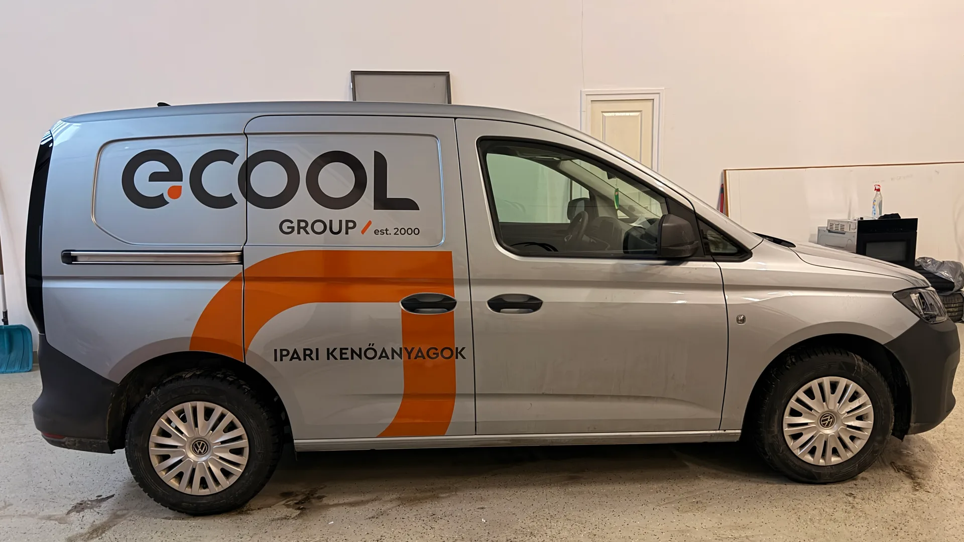 Ecool Volkswagen Caddy oldalnézet – ezüst karosszérián nagyméretű fekete e.COOL logó, GROUP est. 2000 felirat, narancssárga D-grafika és IPARI KENŐANYAGOK felirat