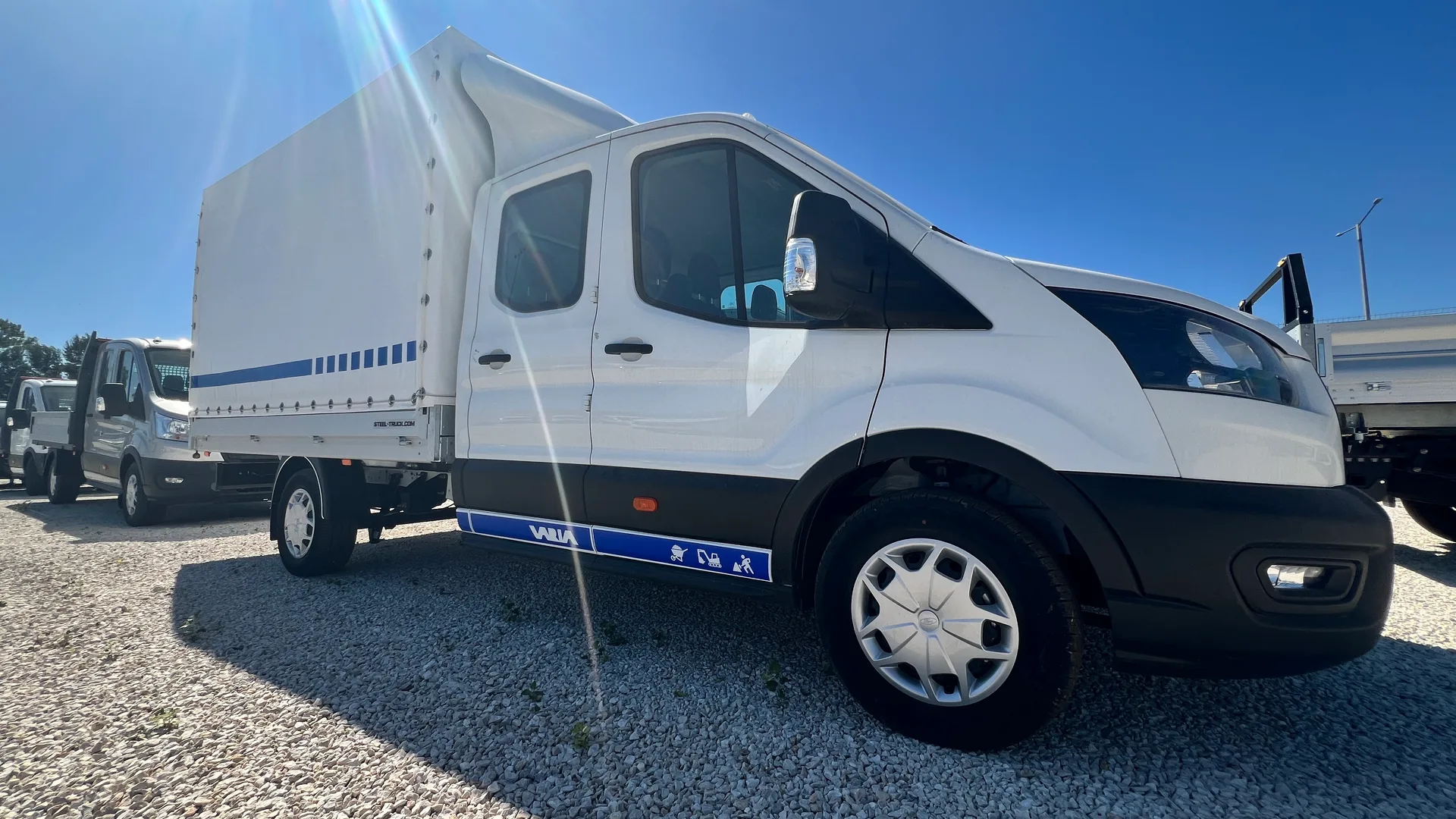 Fehér Ford Transit duplakabin ponyvás felépítménnyel és kék VARIA küszöbdekorral a Ford Solymár telephelyén