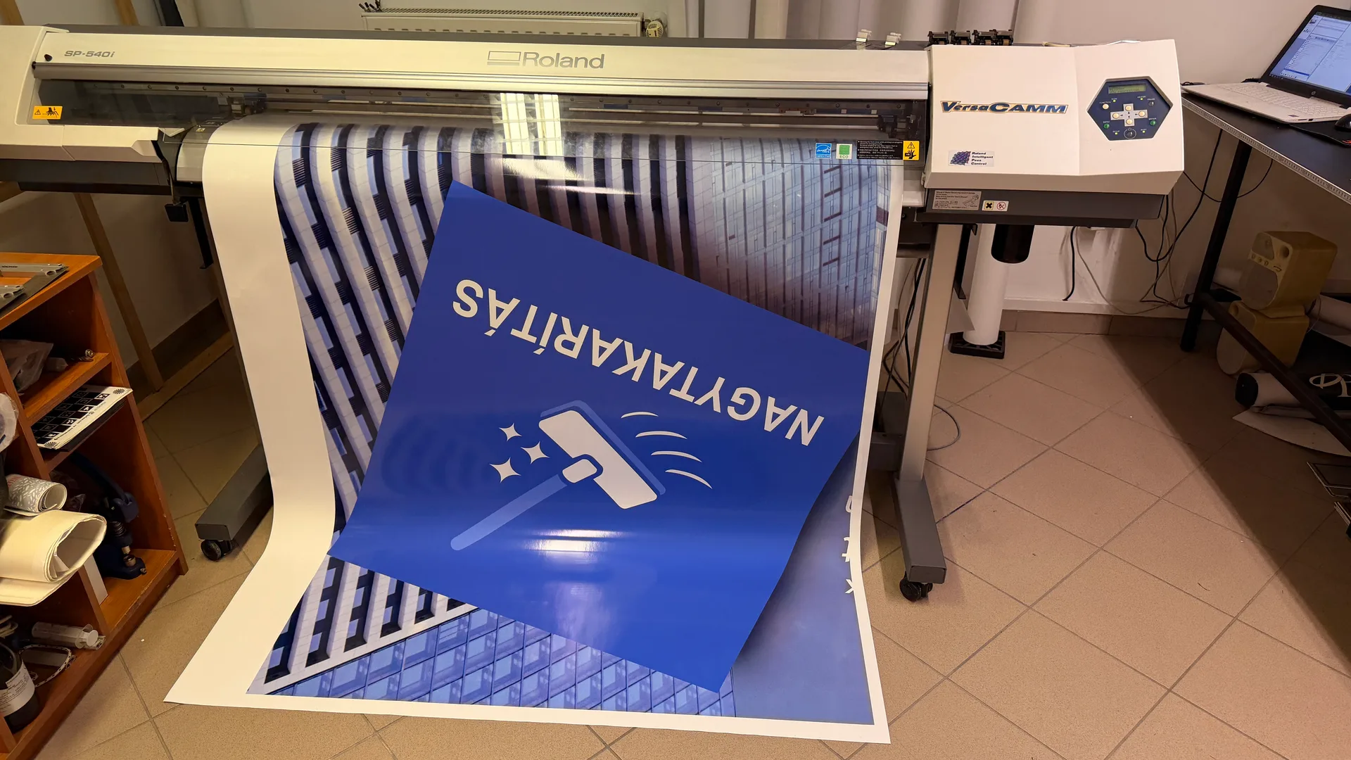 HW Solutions járműdekoráció gyártás – Roland VersaCAMM SP-540i nyomtatóból kifutó épület-homlokzat grafika és nyomtatott NAGYTAKARÍTÁS panel a takarító figurával