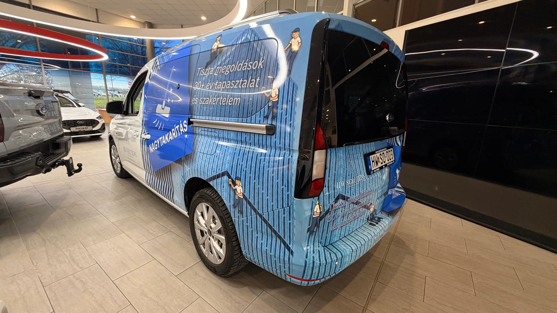 HW Solutions Ford Tourneo Connect hátsó-oldali nézet – épület-homlokzat motívum, illusztrált takarítófigurák, weboldal és kék NAGYTAKARÍTÁS grafika