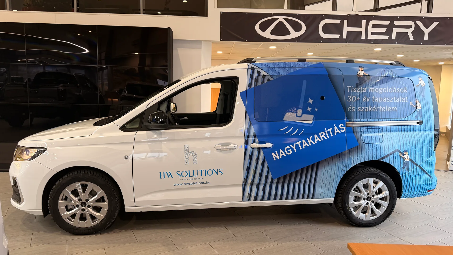 HW Solutions Ford Tourneo Connect bal oldali teljes nézet Chery márkakereskedésben – fehér karosszérián vágott HW Solutions logó és kék épület-homlokzat grafika