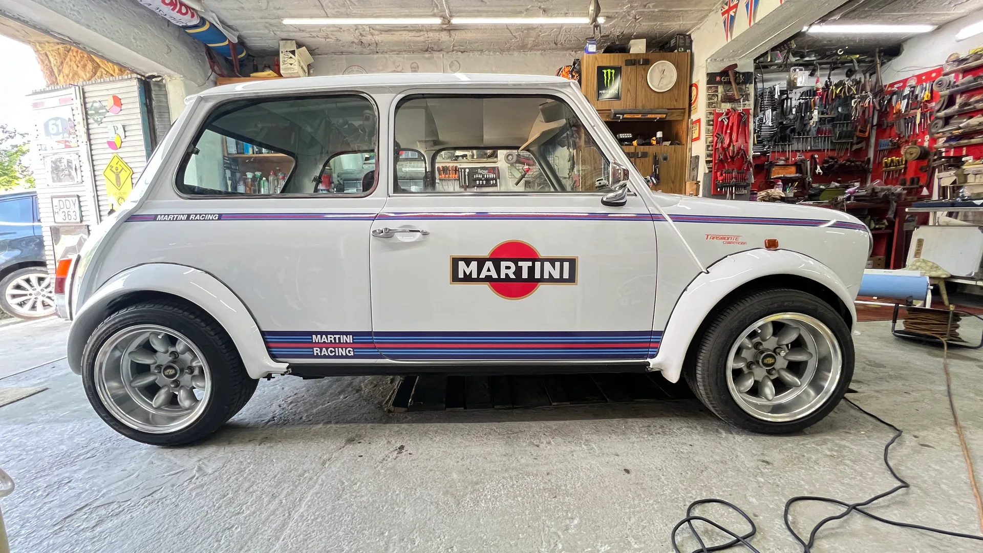 Fehér klasszikus Mini oldalnézetből, kék-piros Martini Racing csíkozással és nagy MARTINI logóval az ajtón
