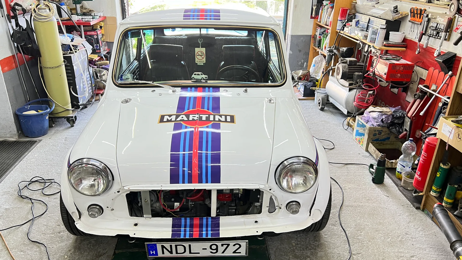 Klasszikus Mini szemből, középen futó Martini Racing csíkozással és MARTINI logóval a motorháztetőn – restaurálási műhelykörnyezetben