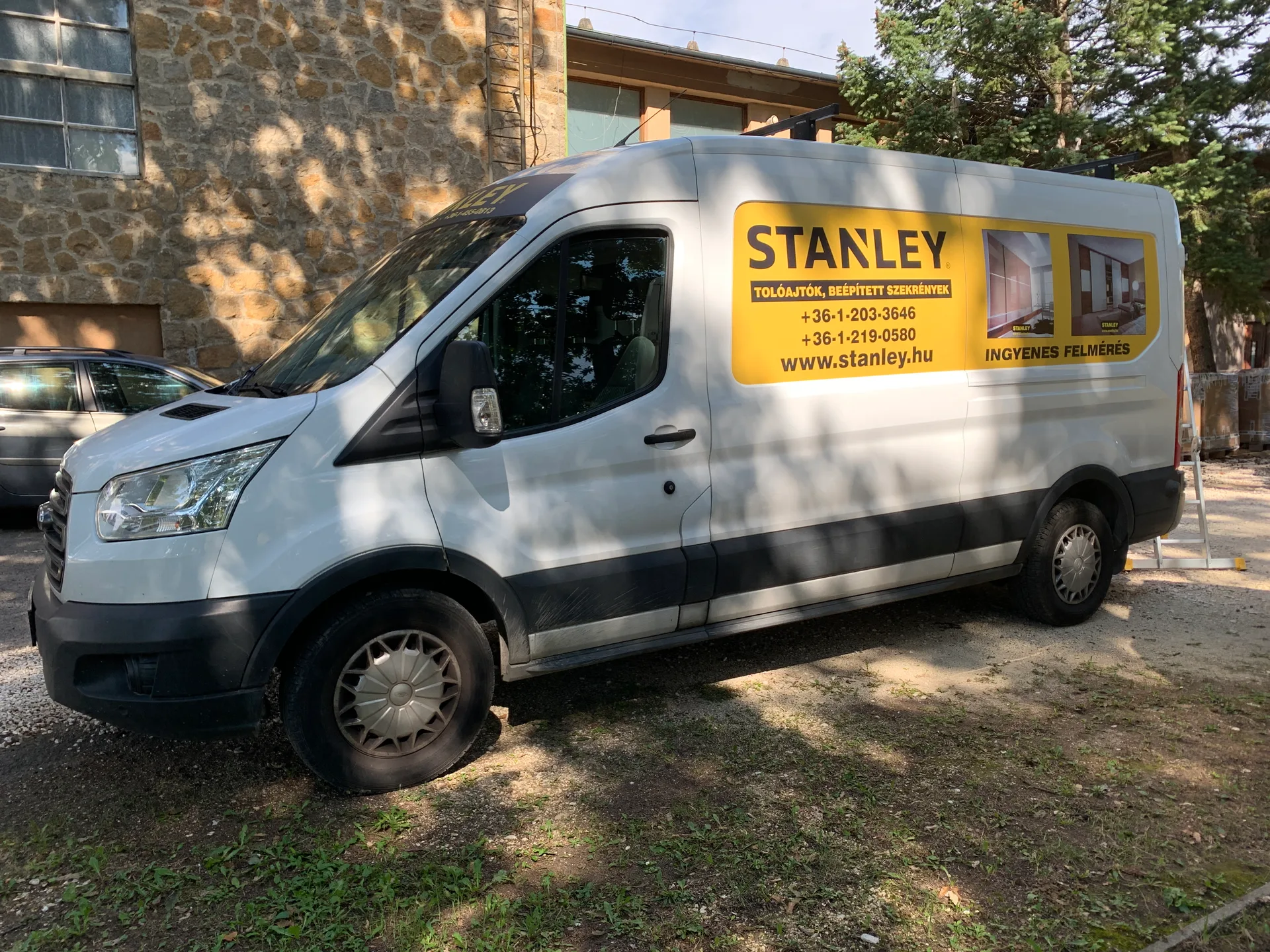Stanley Ford Transit furgon teljes bal oldali nézete – a két hátsó vakablakot lefedő sárga Stanley dekorpanel a logóval, szolgáltatásokkal, telefonszámokkal, weboldalcímmel és INGYENES FELMÉRÉS üzenettel