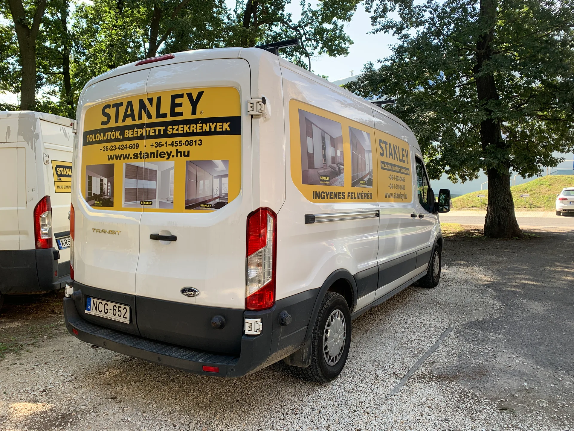 Fehér Stanley Ford Transit furgon hátulsó háromnegyedes jobb oldali nézete – a jobb oldali vakablakon és a hátsó ajtók felső felén sárga Stanley dekor a referenciafotókkal és kontaktokkal