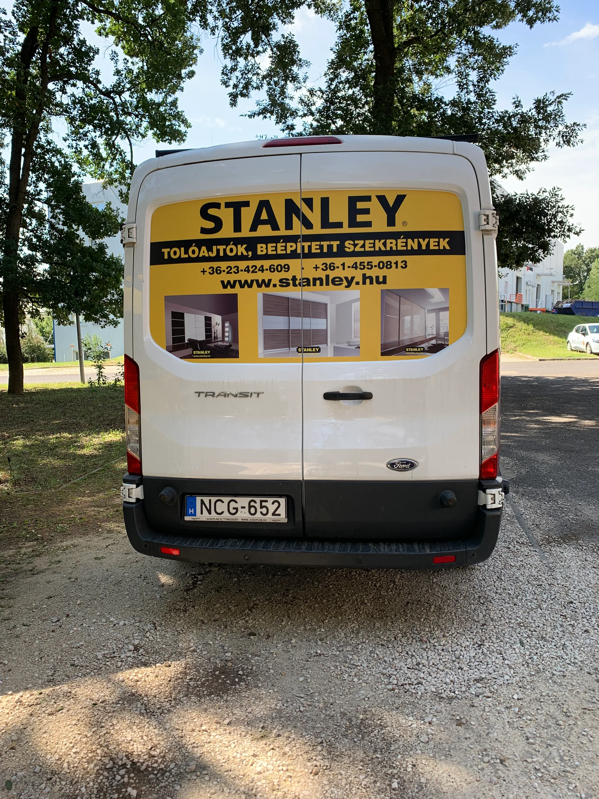 Fehér Stanley Ford Transit furgon hátulnézete – a hátsó ajtók felső részén szimmetrikusan elhelyezett sárga Stanley dekorpanel logóval, telefonszámokkal, weboldalcímmel és beépített szekrényes referenciafotókkal