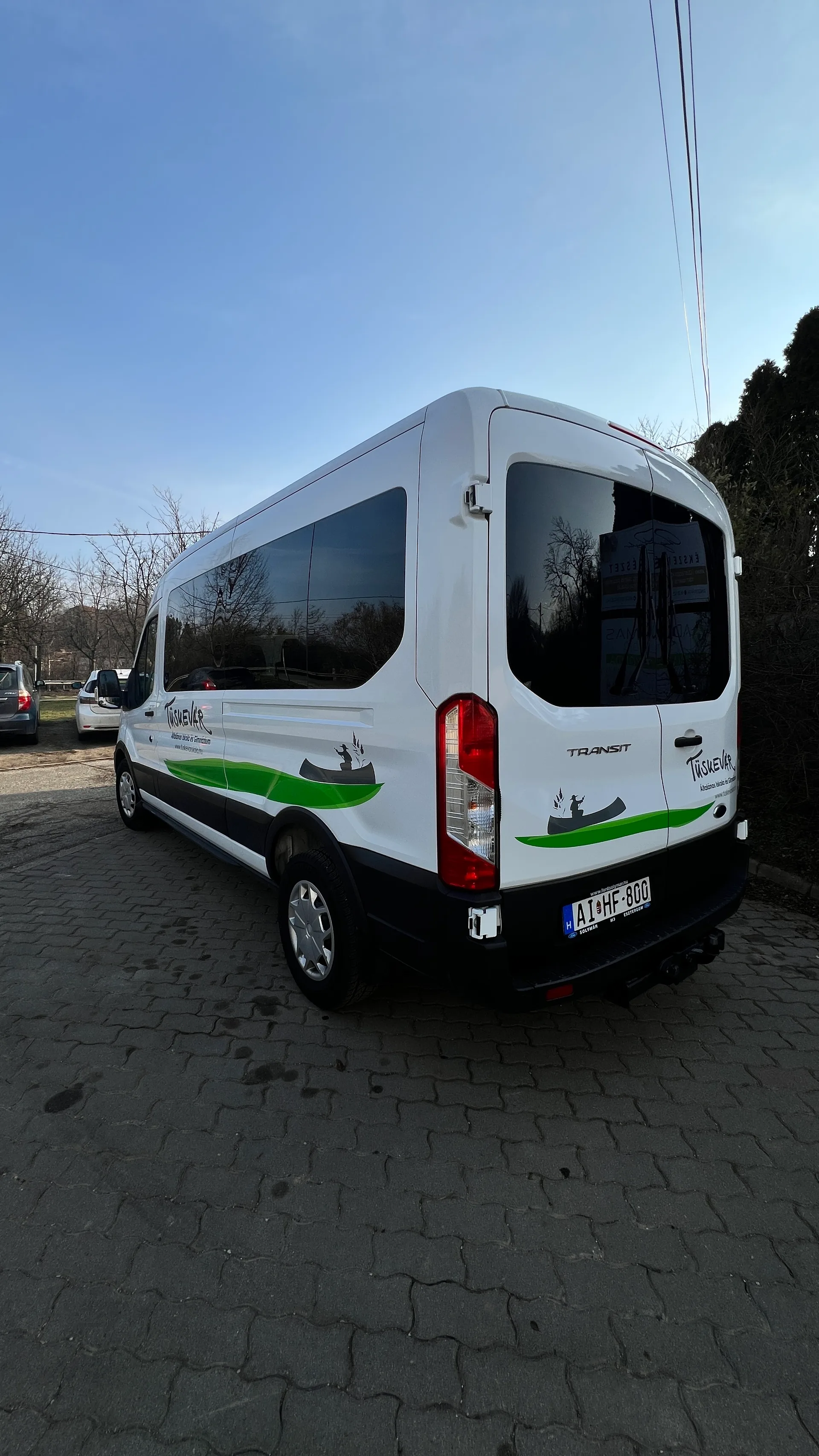 Álló formátumú hátsó-oldalsó fotó a fehér Ford Transit buszról, zöld-szürke arculati elemekkel